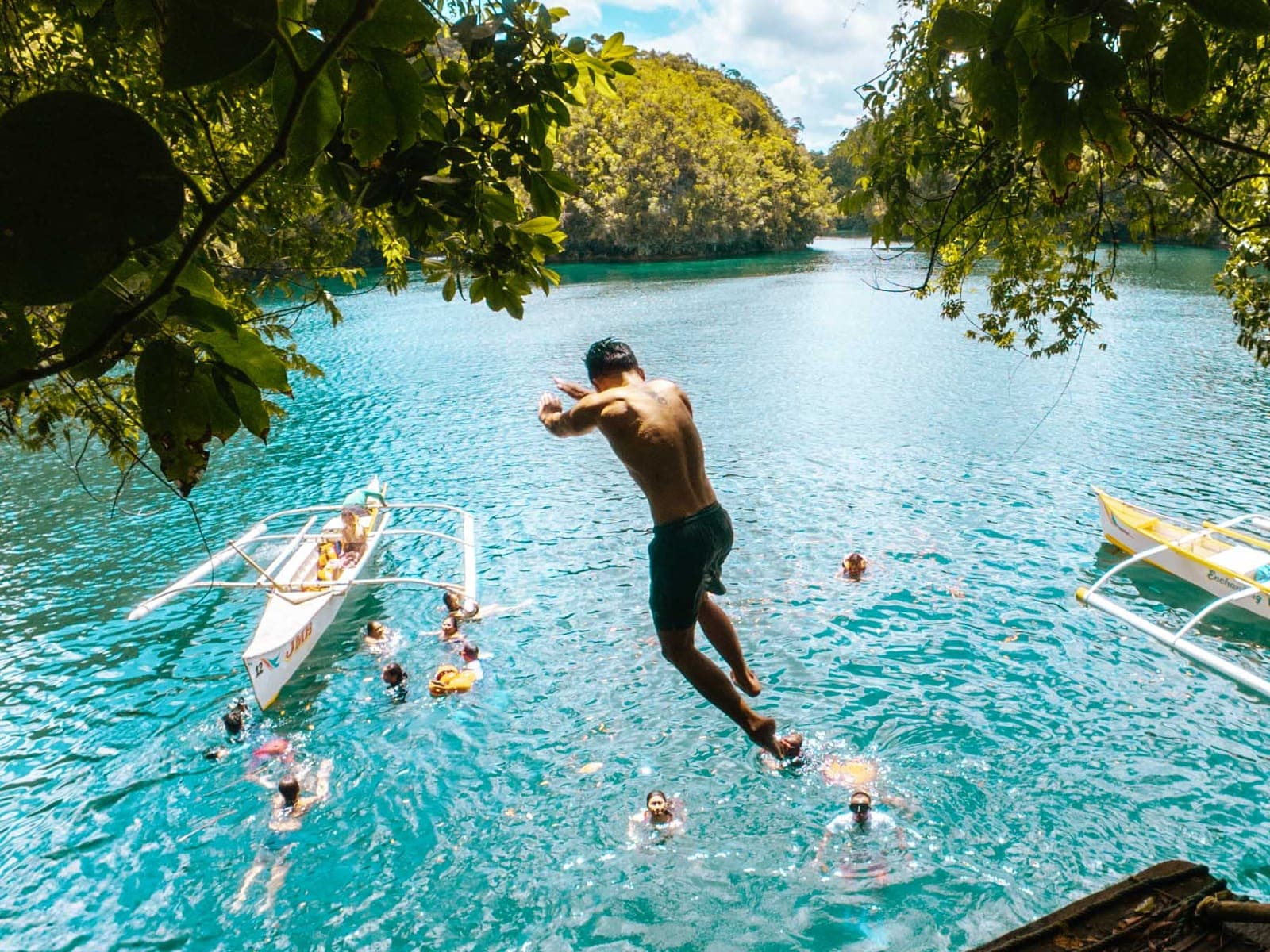 A swimmer jumping from a wooden platform into the turquoise water of Sohoton Cove, with bangka boats and swimmers below